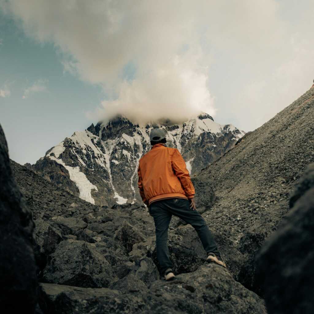 A lone hiker in an orange jacket climbs a rocky mountain in Cusco, Peru, under a cloudy sky.